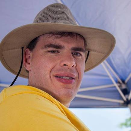 A photo of Matt wearing an adventure hat and a yellow shirt on a sunny day under an umbrella